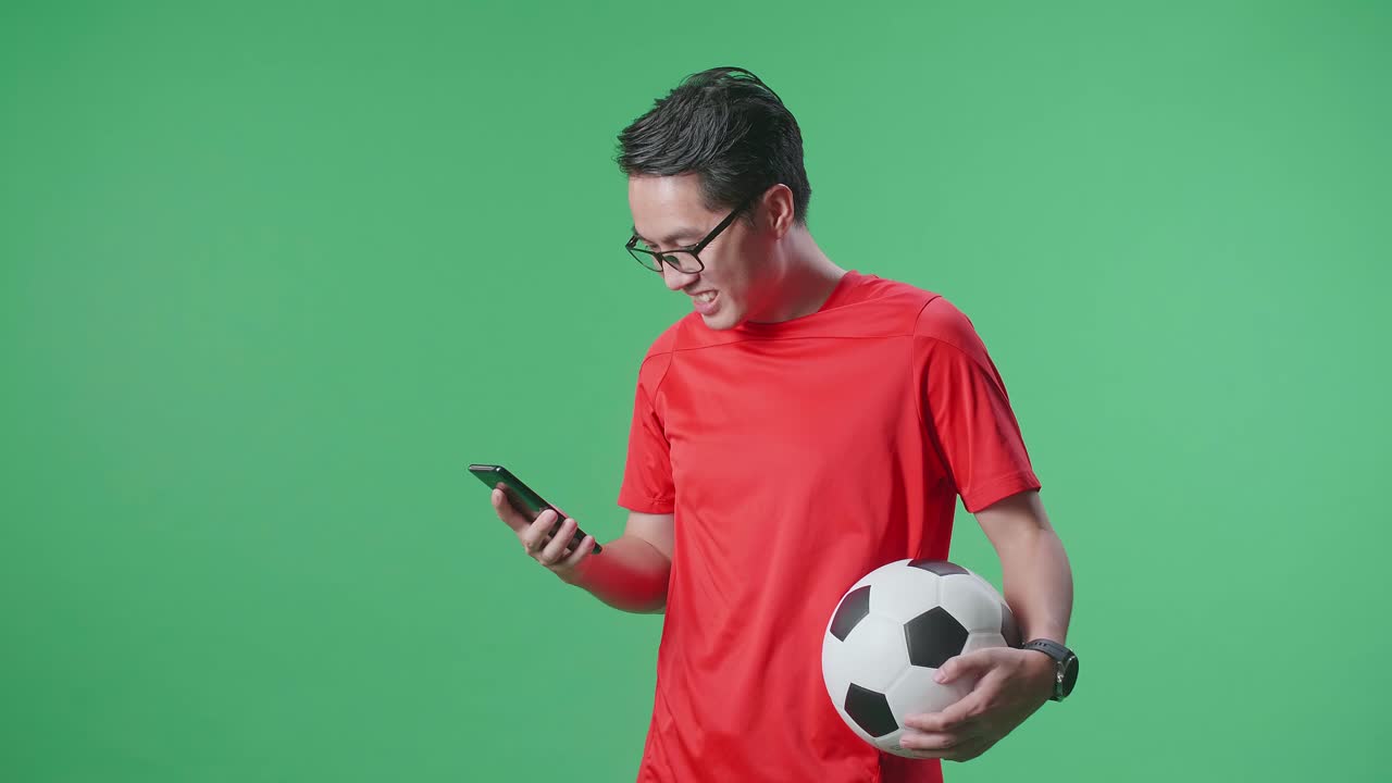 Vista laterale di un uomo asiatico sorridente con un pallone che usa lo smartphone mentre tifa per il calcio su sfondo verde.