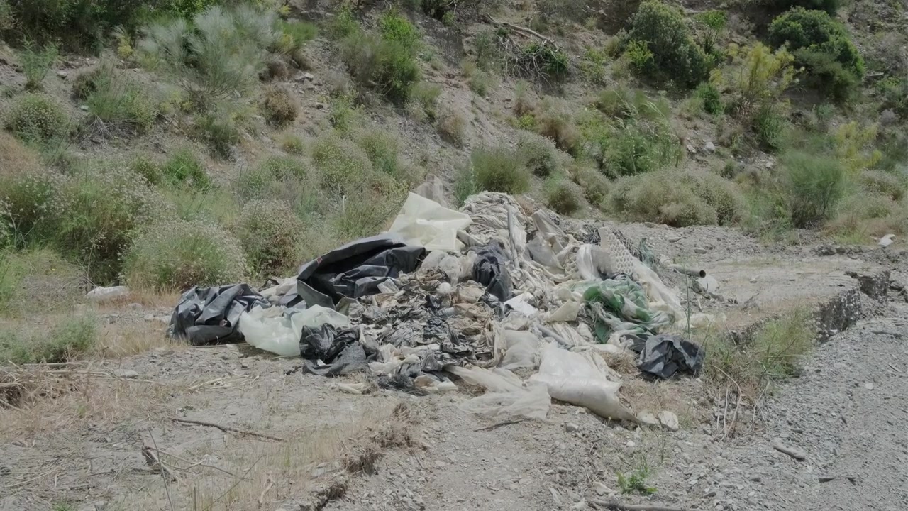 Inquadratura media di sacchi di plastica, teloni e altri tipi di spazzatura provenienti da agricoltura intensiva e serre, abbandonati sul letto di un fiume in Spagna.