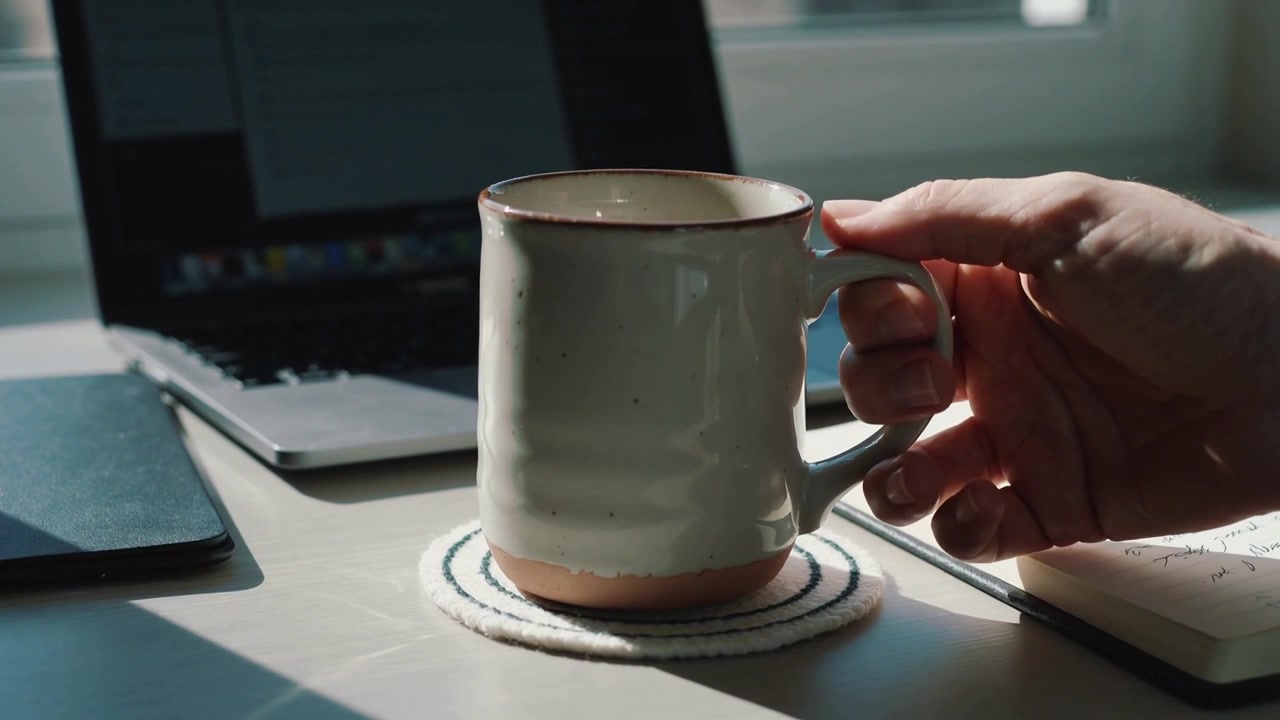Mano destra che entra da destra e posiziona una tazza di ceramica su un sottobicchiere mentre si lavora al laptop