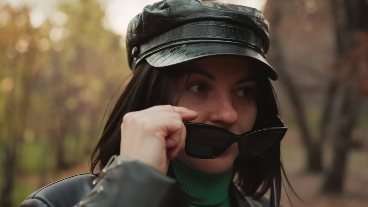 Primo piano di una donna con un berretto che si sistema gli occhiali da sole su un sentiero nel bosco, ritratto intimo con sguardo riflessivo e sorriso accennato, dettagli di dolcevita verde e berretto in pelle lucida, sfondo bokeh leggero e autunnale