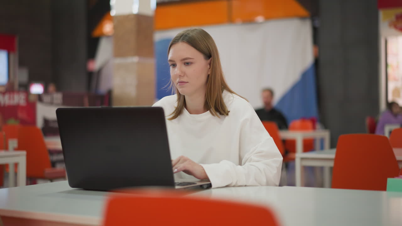 mujer trabajando remotamente en una computadora portátil en un restaurante, mirando momentáneamente hacia otro lado antes de reanudar el trabajo, decoración vibrante y fondo borroso con personas a su alrededor