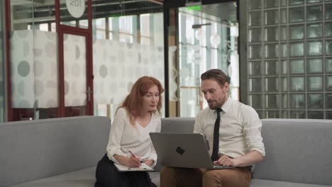 a red haired businesswoman has a relaxed meeting with a young man with a laptop on the couch in the common areas of the office building 1