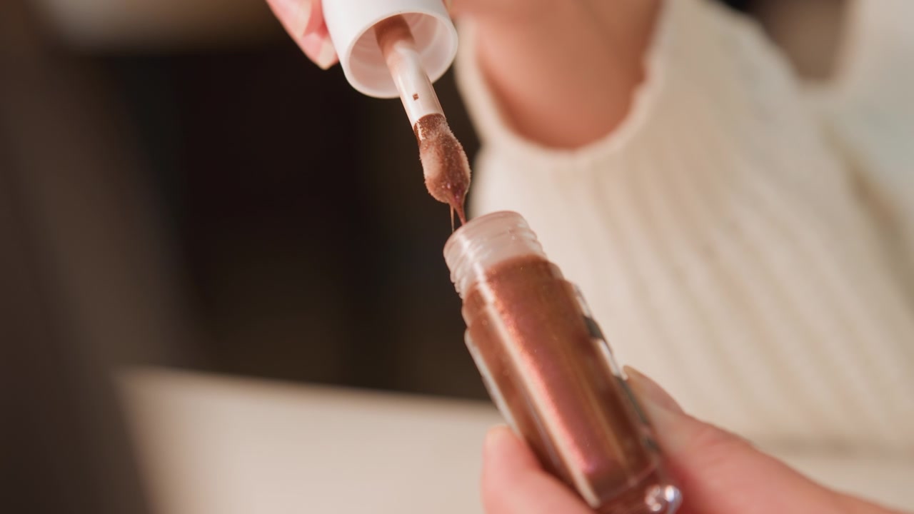 Close-up view of hands holding false nail tip while brush lifts above surface with clear polish dripping from bristles back toward bottle, showing wet shine, careful grooming, and salon style beauty