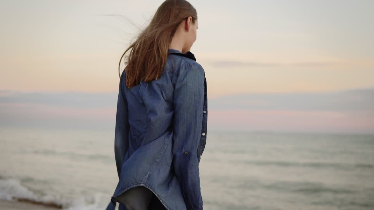 Back view of attractive woman coming up to the sea and trying temperature of water with her hand on the beach during sunset