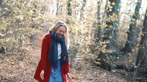 Young-beautiful-woman-throwing-leaves-in-the-air-amidst-thee-orange-brown-autumn-forest-woodland-while-wearing-a-red-coat