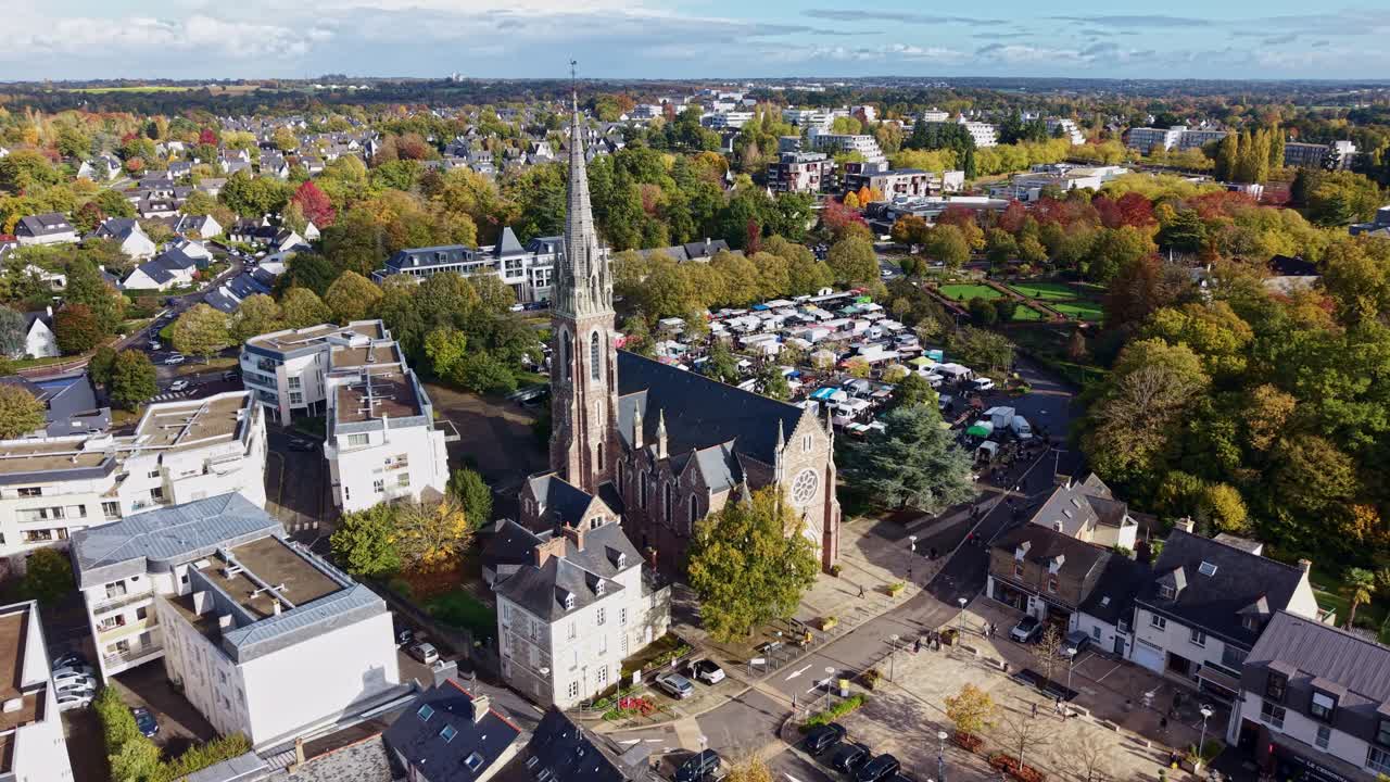 Drone orbiting shot around Saint-Martin Church and the market square in Cesson-Sévigné, showing colorful autumn trees, city buildings, parking, and a clear blue sky