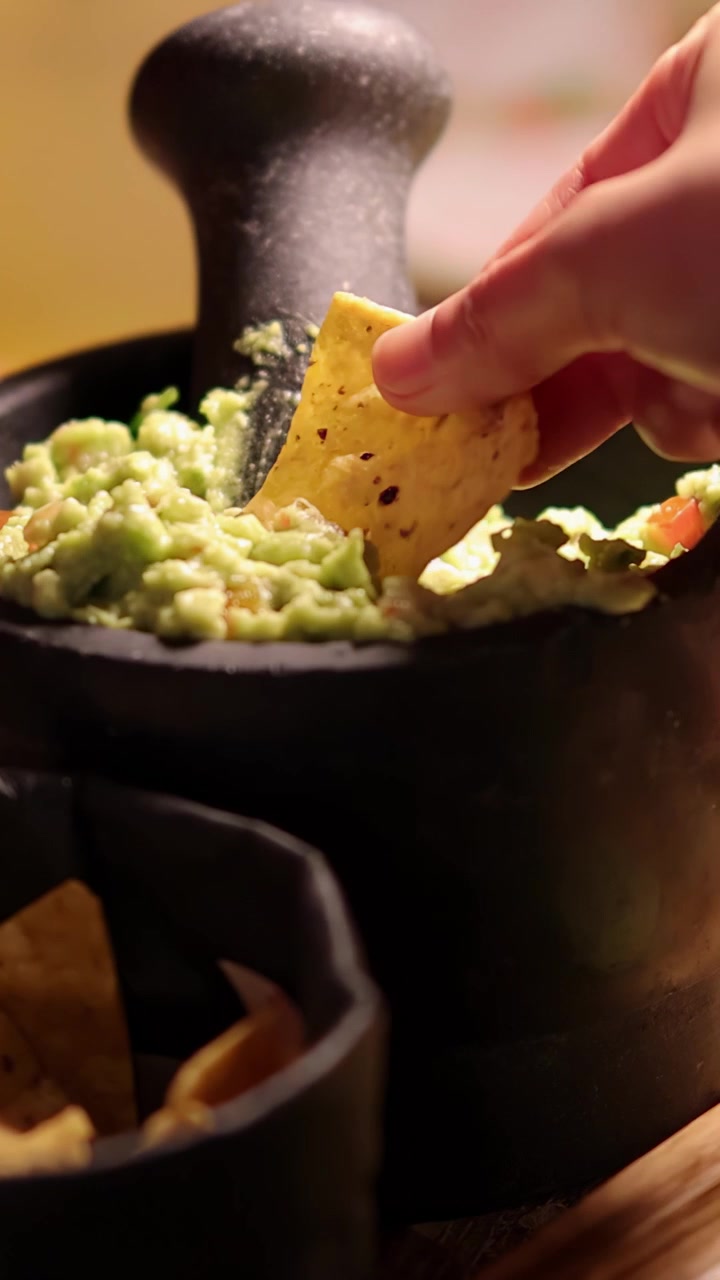 Una mano intinge un nacho nel guacamole denso servito in un tradizionale molcajete di pietra, sotto un'illuminazione calda e soffusa con una profondità di campo ridotta.