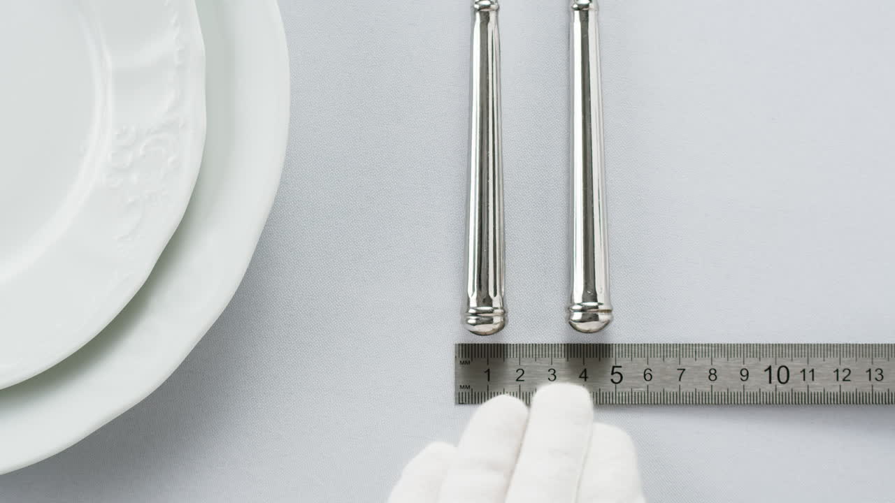 Minimalist table setting with white plate, fork, and knife arranged neatly, and a gloved hand adjusting cutlery.