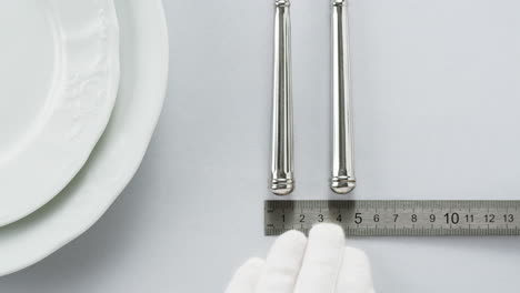 minimalist table setting with white plate, fork, and knife arranged neatly, and a gloved hand adjusting cutlery.