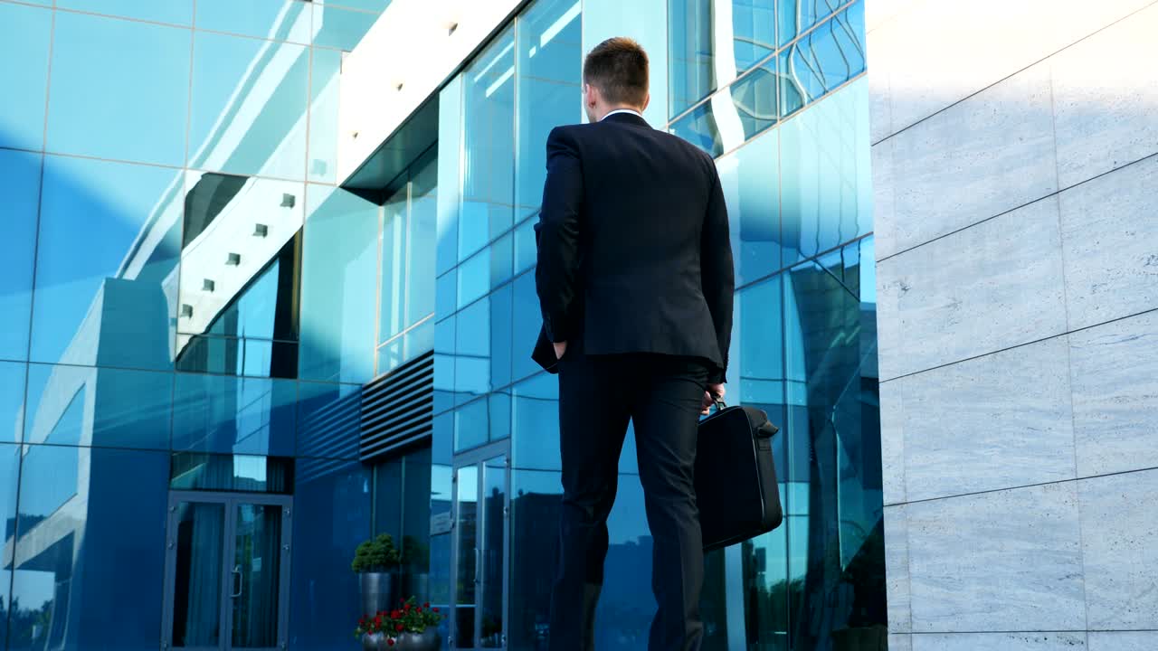 Giovane uomo d'affari che si reca al lavoro. Uomo sicuro di sé in abito in viaggio verso l'ufficio. Uomo d'affari con una ventiquattrore che cammina per strada in città vicino a un edificio moderno. Rallentatore. Vista posteriore. Primo piano.