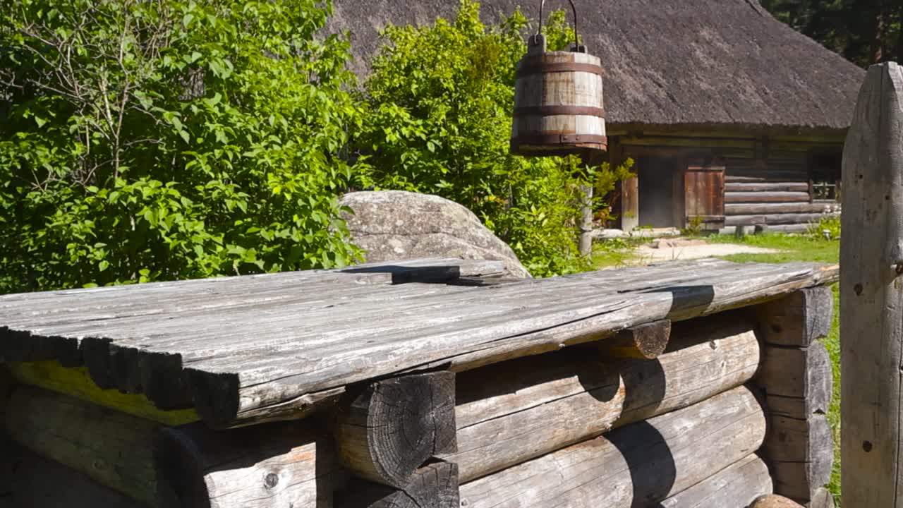 Old traditional wooden brown vintage well with a wooden bucket with rusty straps hanging in front of a old traditional village log cabin with a thatched roof during summer sunny day in a green garden
