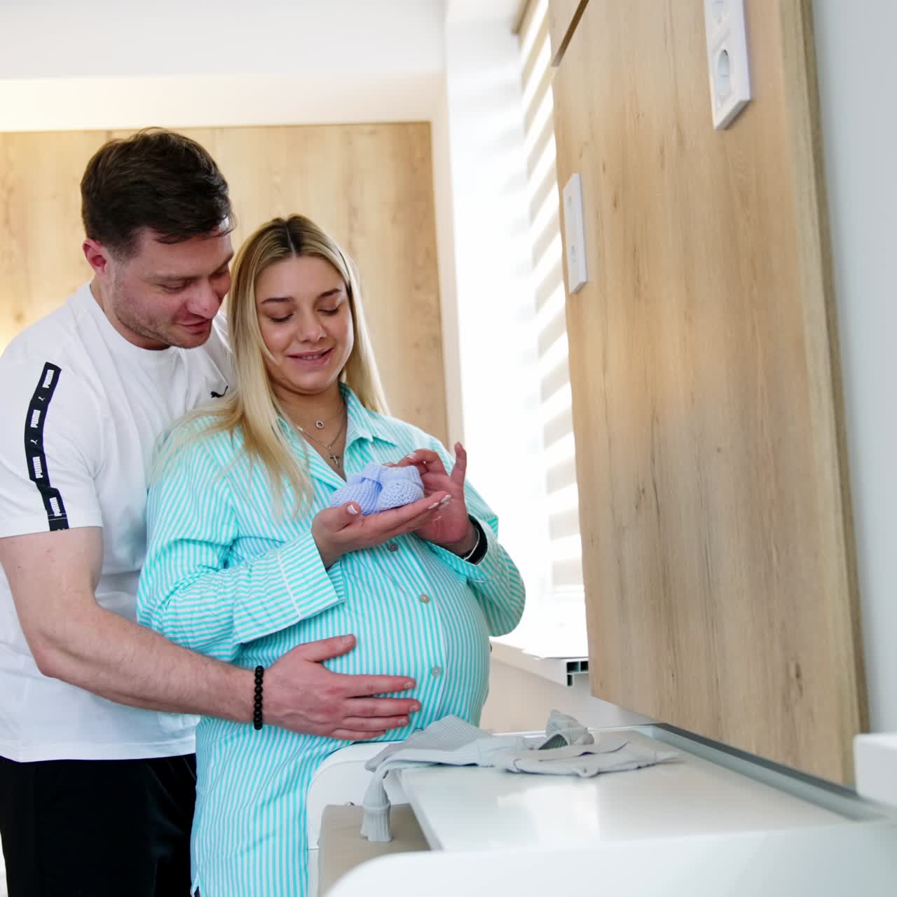 Happy expecting couple stand in hospital ward hugging. Woman holds the tiny socks for baby and man strokes her belly