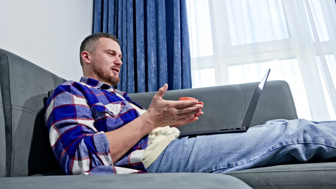 En mand sidder på sofaen med en bærbar computer, mens han holder en kop og fører en samtale derhjemme om dagen