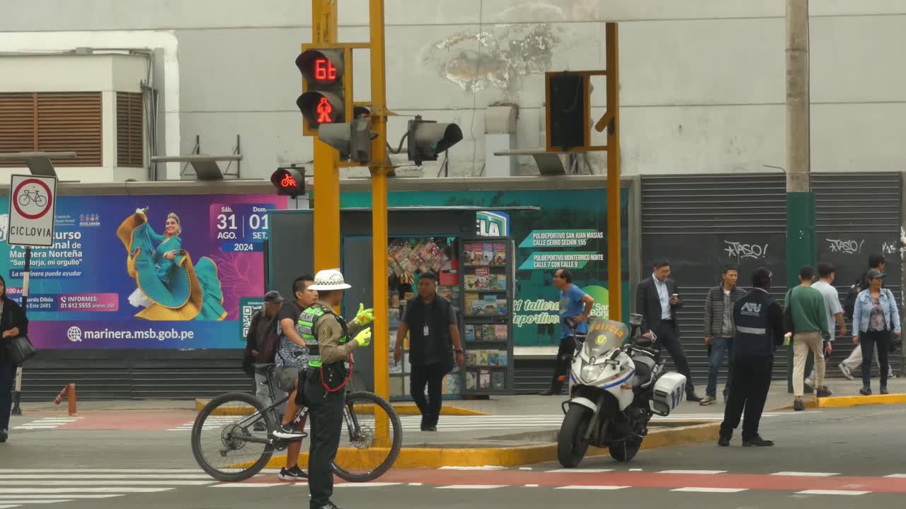 A Peruvian traffic police directing traffic flow. Several people walk by in background. A police car drives by.