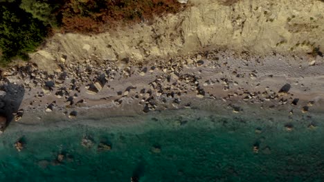 Felsiger-Strand-Mit-Sand-Und-Klippen,-Die-Zur-Goldenen-Stunde-Im-Mittelmeer-Von-Smaragdgrünem-Sauberem-Meerwasser-Gewaschen-Werden,-Von-Oben-Nach-Unten-Aus-Der-Luft