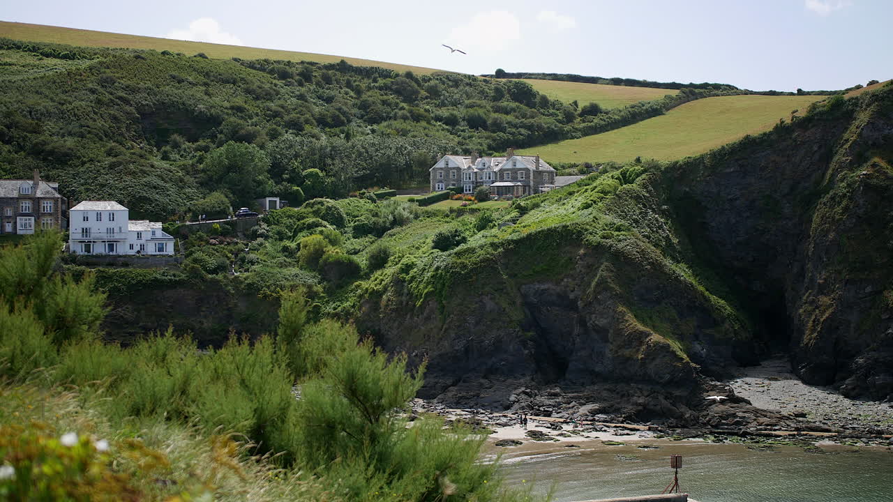 Houses are perched on green hills above rocky cliffs in Cornwall, showing the real-life village of Port Isaac known as Portwenn