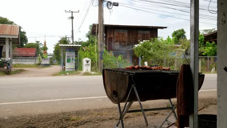 street vendor cooking kai yang chicken on grill as vehicle traffic passes, thailand