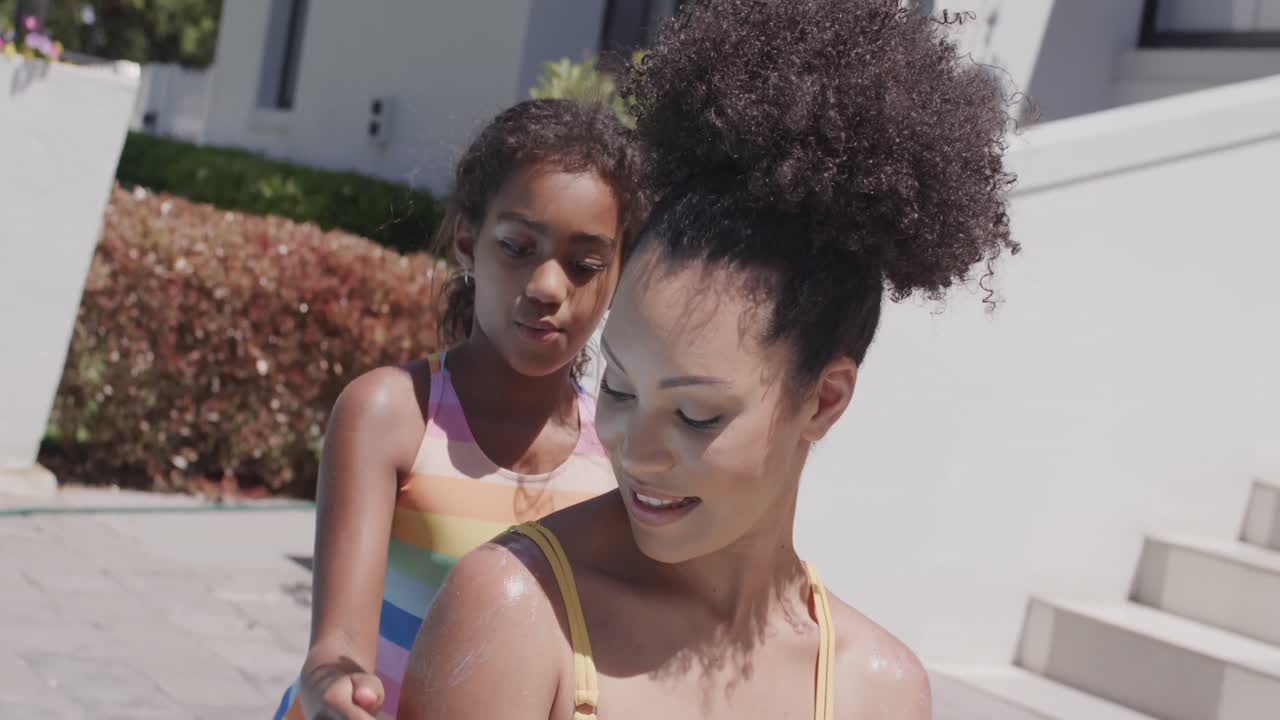 Happy african american mother and daughter applying sunscreen by swimming pool, slow motion