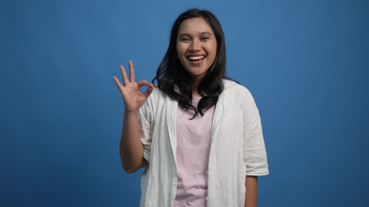 young asian woman 20s years old posing isolated on blue background studio. People lifestyle concept. Looking camera say wow showing OK gesture blinking