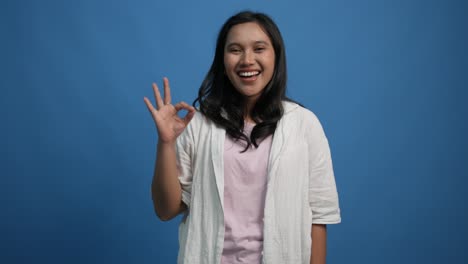 young asian woman 20s years old posing isolated on blue background studio. people lifestyle concept. looking camera say wow showing ok gesture blinking