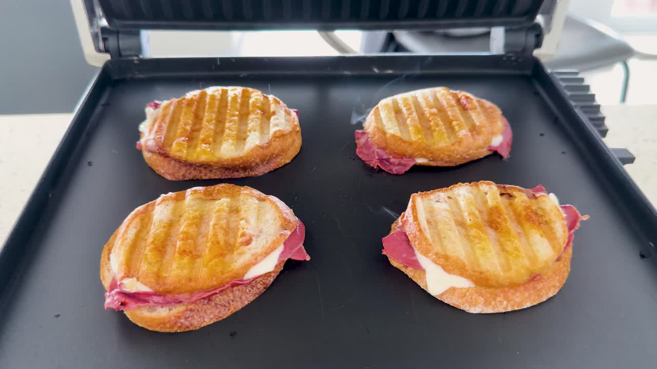 Inquadratura statica dall'alto di quattro sandwich che tostano su una piastra elettrica per panini, con vapore che sale.