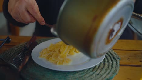 a close-up of a hand spooning cooked farfalle pasta onto a white plate from a saucepan on a rustic wooden table. daugavpils, latvia (latgale)