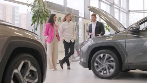 young car salesman showing to young couple new automobile at dealership salon.