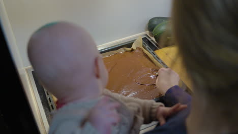 mother with newborn baby spreading brownie dough on baking dish tray at kitchen