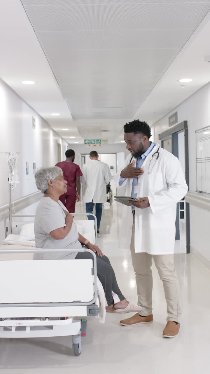 Vertical video of diverse male doctor with tablet talking to senior ...