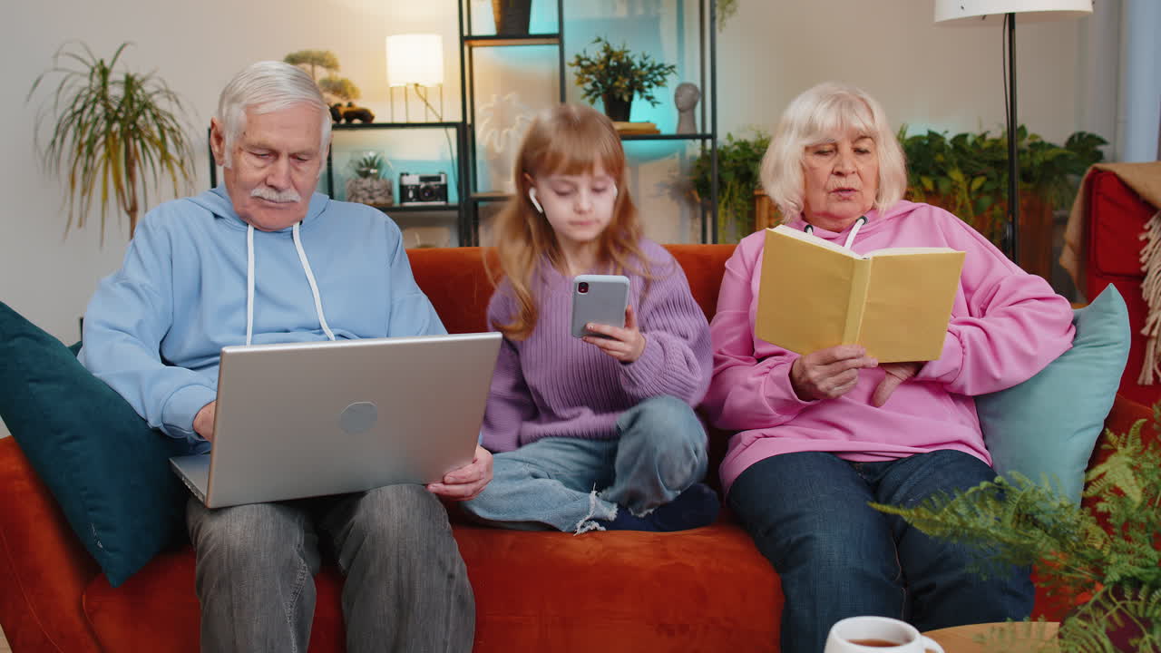 Girl granddaughter listening to music sitting amidst grandparents using laptop and reading book