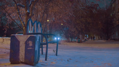 empty bench on a snowy path illuminated by soft lights with blurred figures in the background, passing cars with glowing headlights, and serene winter atmosphere under snow-laden trees