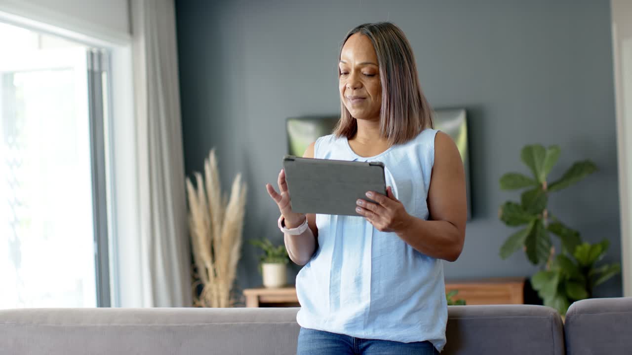 Donna afroamericana che usa un tablet sul divano di casa, guardando una finestra luminosa mentre legge lo schermo.