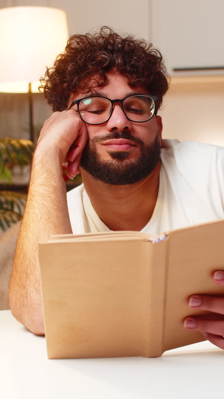 Homem árabe lê livro monótono à noite, apoia-se na mão, entediado e cansado na secretária, em um ambiente de trabalho noturno tranquilo.