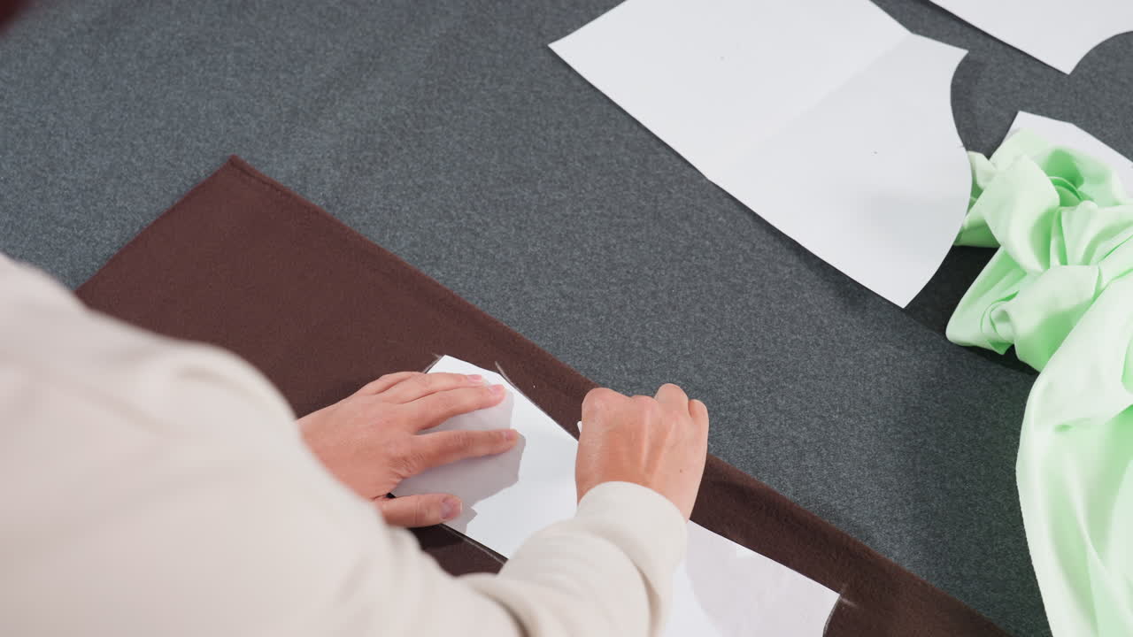 Garment designer sketching pattern with pencil on white paper sheet placed over dark brown fabric while working carefully in tailoring workshop surrounded by scattered sewing materials