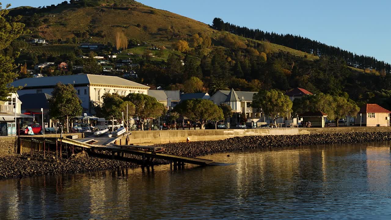 Calm waters and golden light illuminate Akaroa's picturesque waterfront, capturing a tranquil evening scene with gentle camera panning