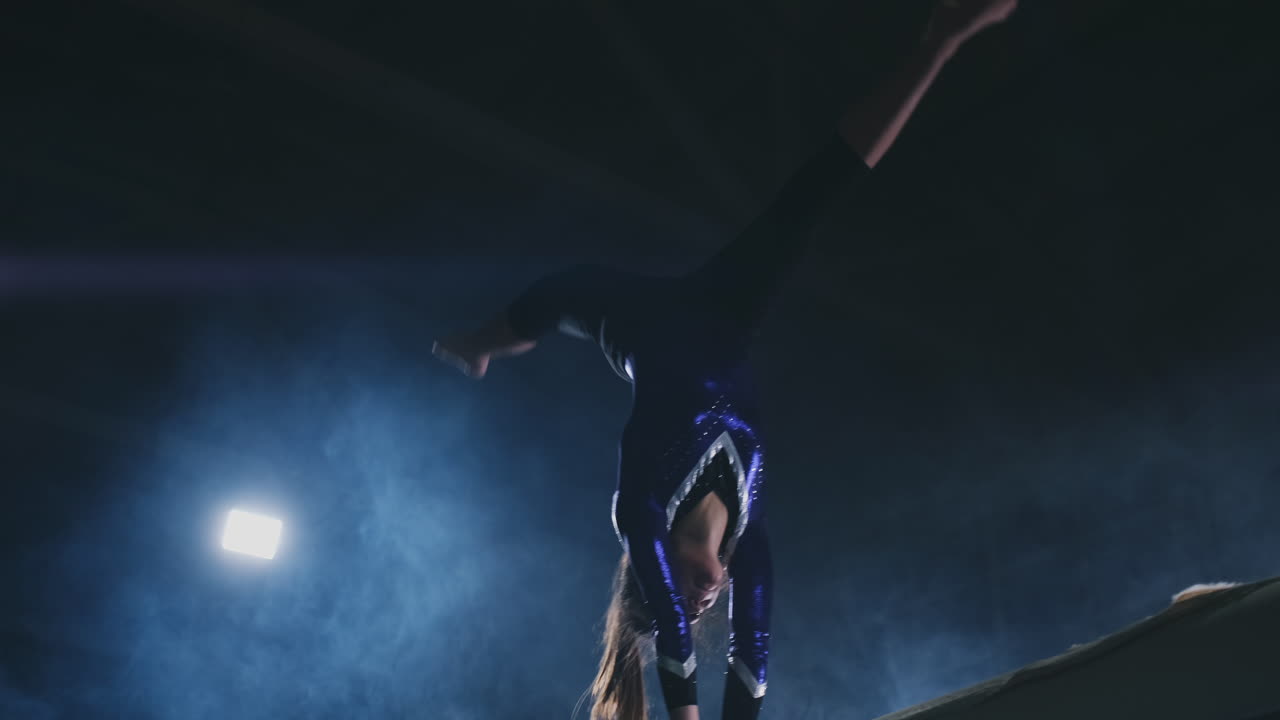 la niña es una atleta profesional realiza un truco acrobático de gimnasia en una viga con luz de fondo y cámara lenta en ropa de gimnasia deportiva. humo y azul. saltar y girar en la viga de equilibrio