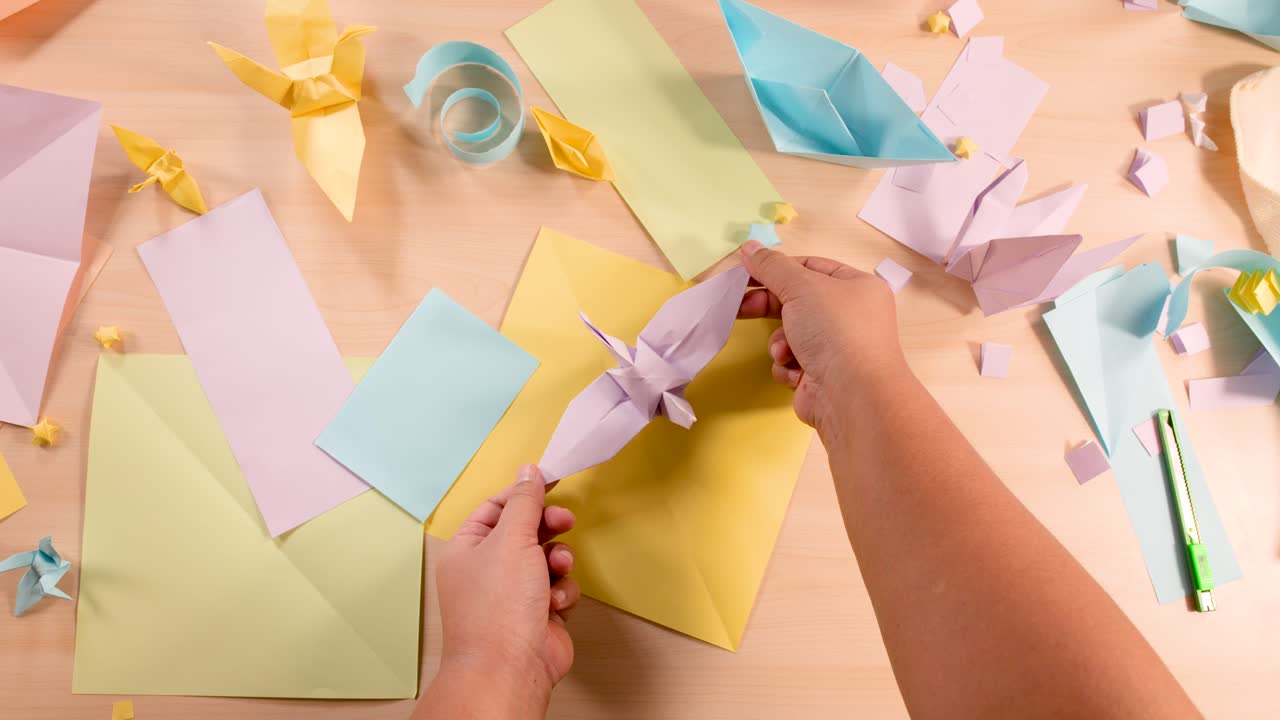 Person folds pastel paper into origami bird, surrounded by colorful sheets, under soft overhead lighting