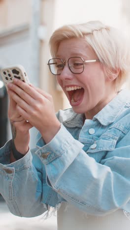 feliz mujer rubia caucásica usando teléfono inteligente celebrando ganar buenas noticias mensaje en la ciudad calle de la ciudad