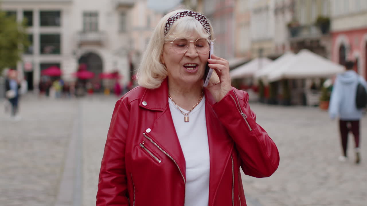 Happy senior woman having remote conversation talking on smartphone good news gossip in city street