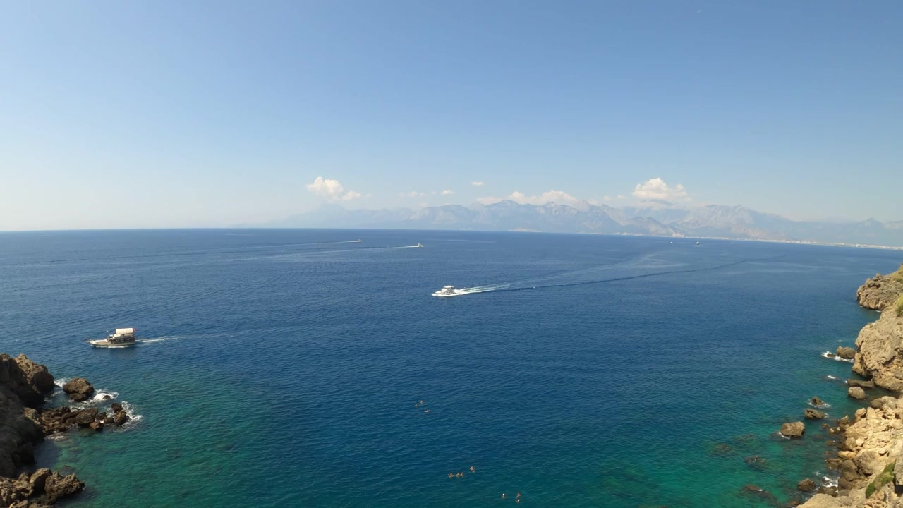 Suggestivo paesaggio marino panoramico mediterraneo con barche, scogliere rocciose e montagne distanti sotto un cielo azzurro limpido lungo la costa di Antalya in Turchia in una giornata di sole.