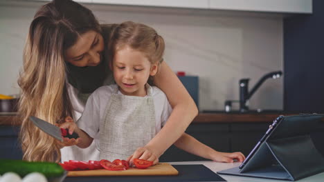 smiling mother and girl in aprons cut fresh tomato on board