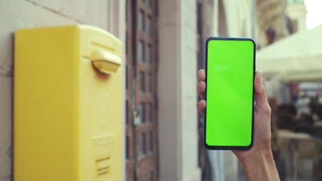 hand holding a green screen smartphone next to a yellow mailbox in an urban setting