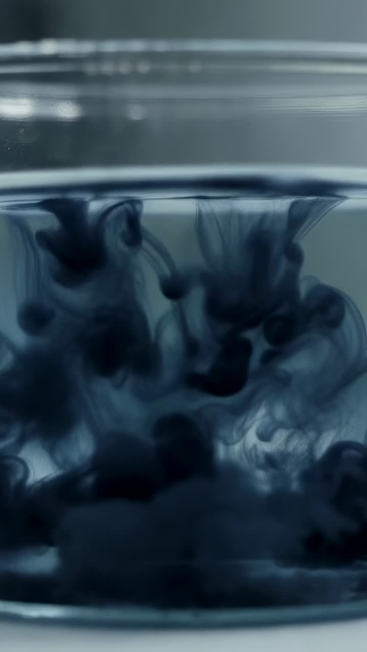 Vertical video: Dropping dark ink sinking in water in glass bowl forming plumes on white lab table