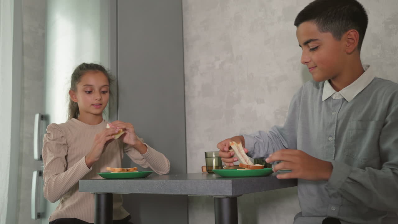 Woman serving food to children, one child looking uninterested, others sitting at table in casual morning kitchen scene, family breakfast time, soft light from window, homey atmosphere