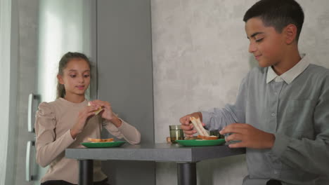 woman serving food to children, one child looking uninterested, others sitting at table in casual morning kitchen scene, family breakfast time, soft light from window, homey atmosphere