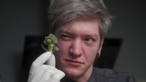 junger verrückter männlicher koch in einem eleganten schwarzen hemd mit alternativem look, tätowierungen, die pilze in der hand halten und exzessiv auf brokkoli in der hand schauen