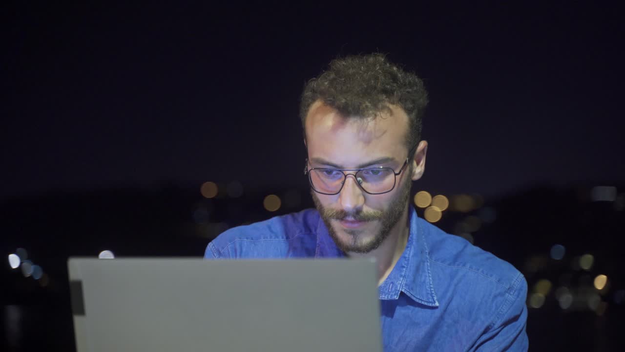 Man working intensely on laptop at night by the sea.
