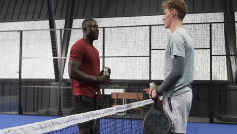 two diverse men enjoying padel tennis match, smiling and holding rackets on indoor court