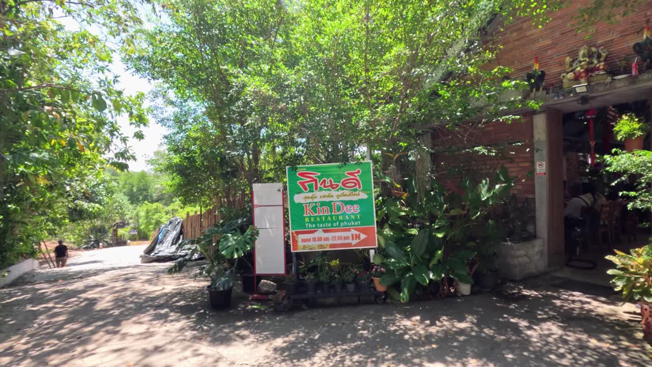 Camera moves toward leafy restaurant entrance, bright daylight, tropical plants, inviting outdoor setting