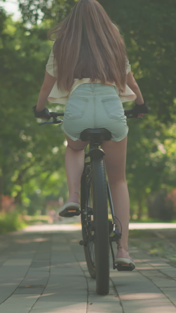 풍경이 아름다운 푸른 공원 길을 자전거를 타고 자신감 있게 달리는 젊은 여성의 뒷모습, 긴 머리카락이 자연스럽게 움직이고, 빛이 나무를 통해 필터링하여 부드러운 그림자를 던집니다.